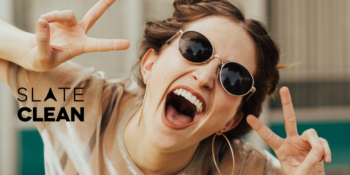 Young woman wearing sunglasses smiling are holding up peace signs with her fingers. Slate Clean logo included.