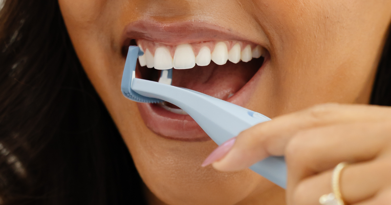 Close up image of a woman with dark hair smiling and using the Slate Origin flosser between her teeth