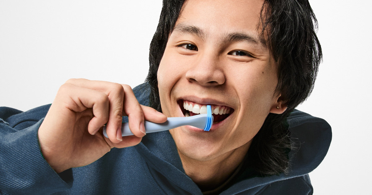 Asian man wearing a hoodie, using a blue Slate mini flosser and smiling. 